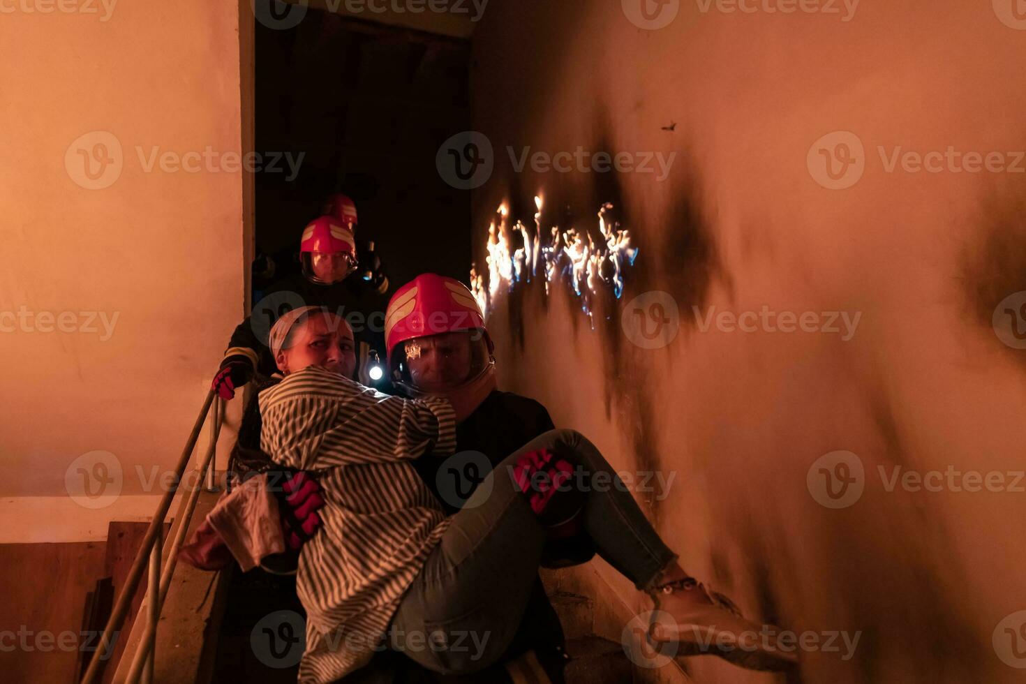 Brave Fireman Descends Stairs of a Burning Building and Holds Saved Girl in His Arms. Open fire and one Firefighter in the Background. photo