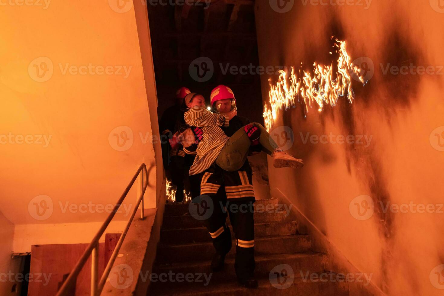 Brave Fireman Descends Stairs of a Burning Building and Holds Saved Girl in His Arms. Open fire and one Firefighter in the Background. photo