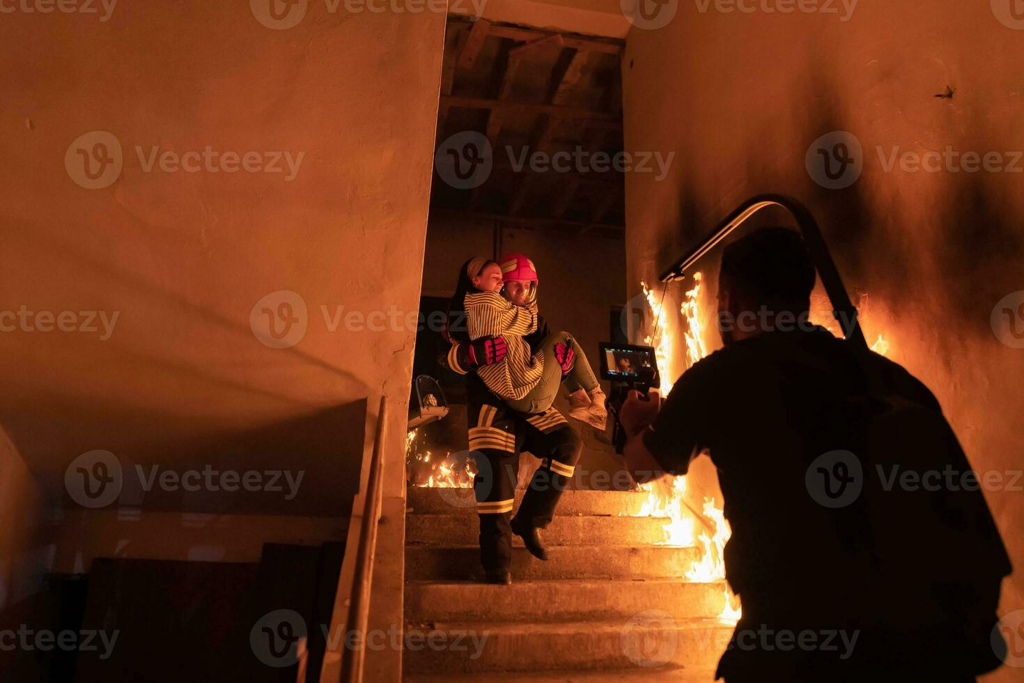 Brave Fireman Descends Stairs of a Burning Building and Holds Saved Girl in His Arms. Open fire and one Firefighter in the Background. photo