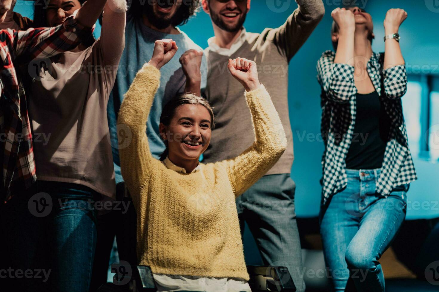 Photo of business women in wheelchairs with their hands raised in the air with their colleagues, together celebrating business success