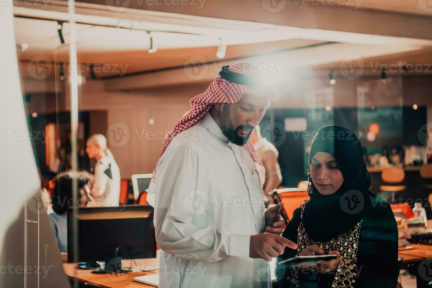 Portrait photo of Arab leader businessman with muslim hijab woman discussing business projects while using tablets and smartphone in modern glass office