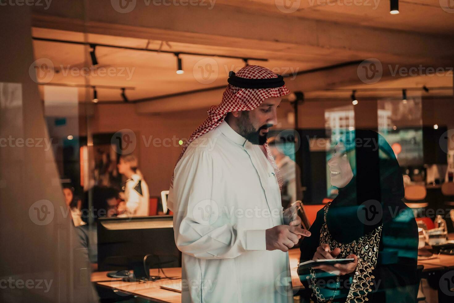 Portrait photo of Arab leader businessman with muslim hijab woman discussing business projects while using tablets and smartphone in modern glass office
