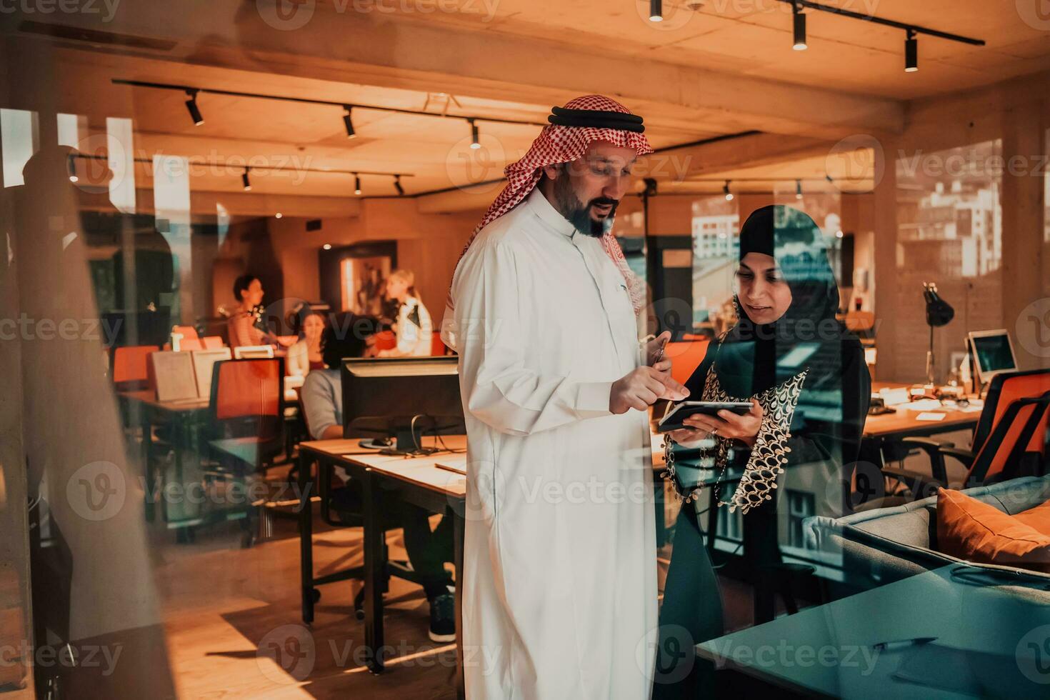 Portrait photo of Arab leader businessman with muslim hijab woman discussing business projects while using tablets and smartphone in modern glass office
