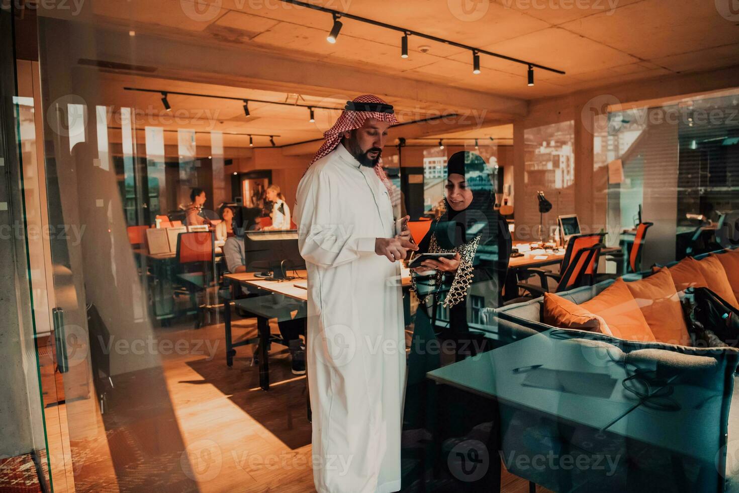Portrait photo of Arab leader businessman with muslim hijab woman discussing business projects while using tablets and smartphone in modern glass office
