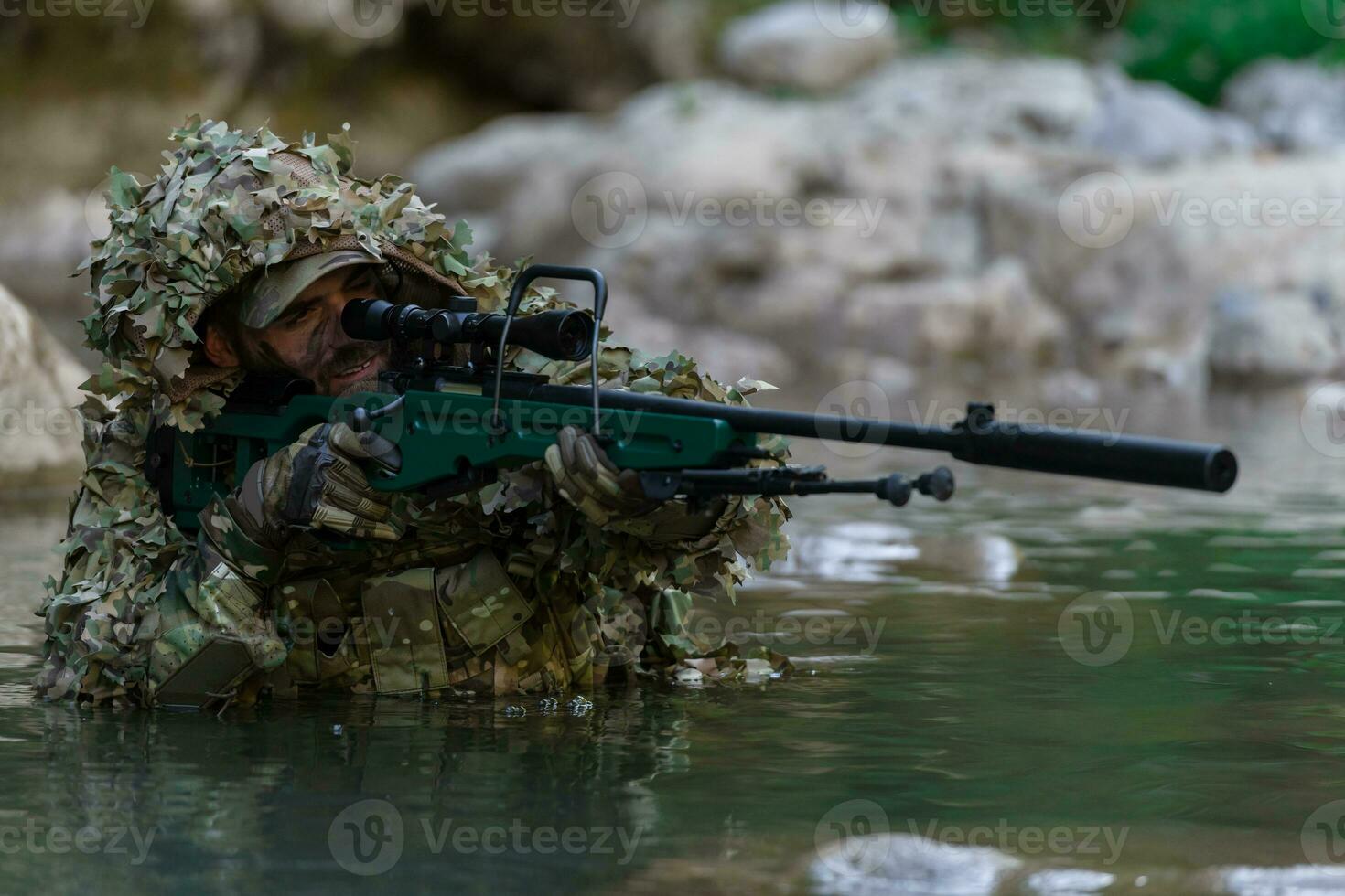 A military man or airsoft player in a camouflage suit sneaking the river and aims from a sniper rifle to the side or to target. photo