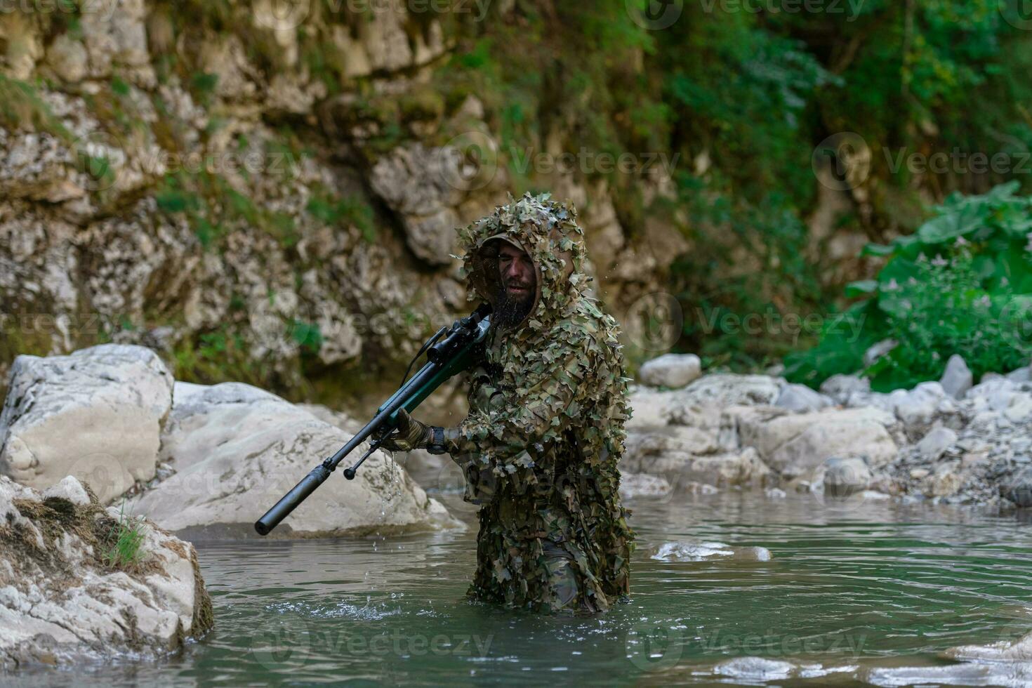 A military man or airsoft player in a camouflage suit sneaking the river and aims from a sniper rifle to the side or to target. photo