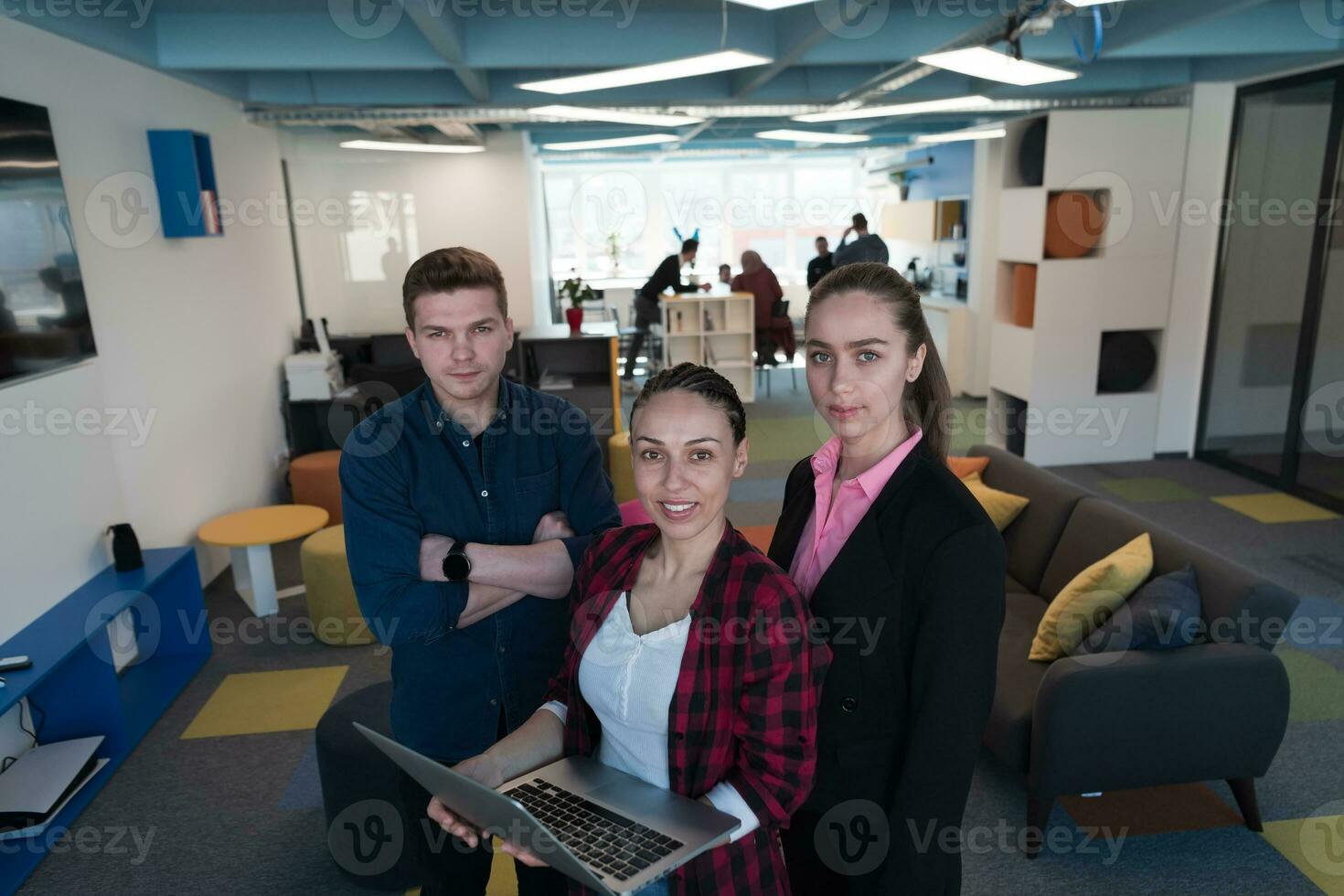 un grupo de joven entusiasta joven negocio personas en moderno oficina. un negocio mujer con un ordenador portátil en su mano, en el antecedentes un colegas con cruzado brazos foto