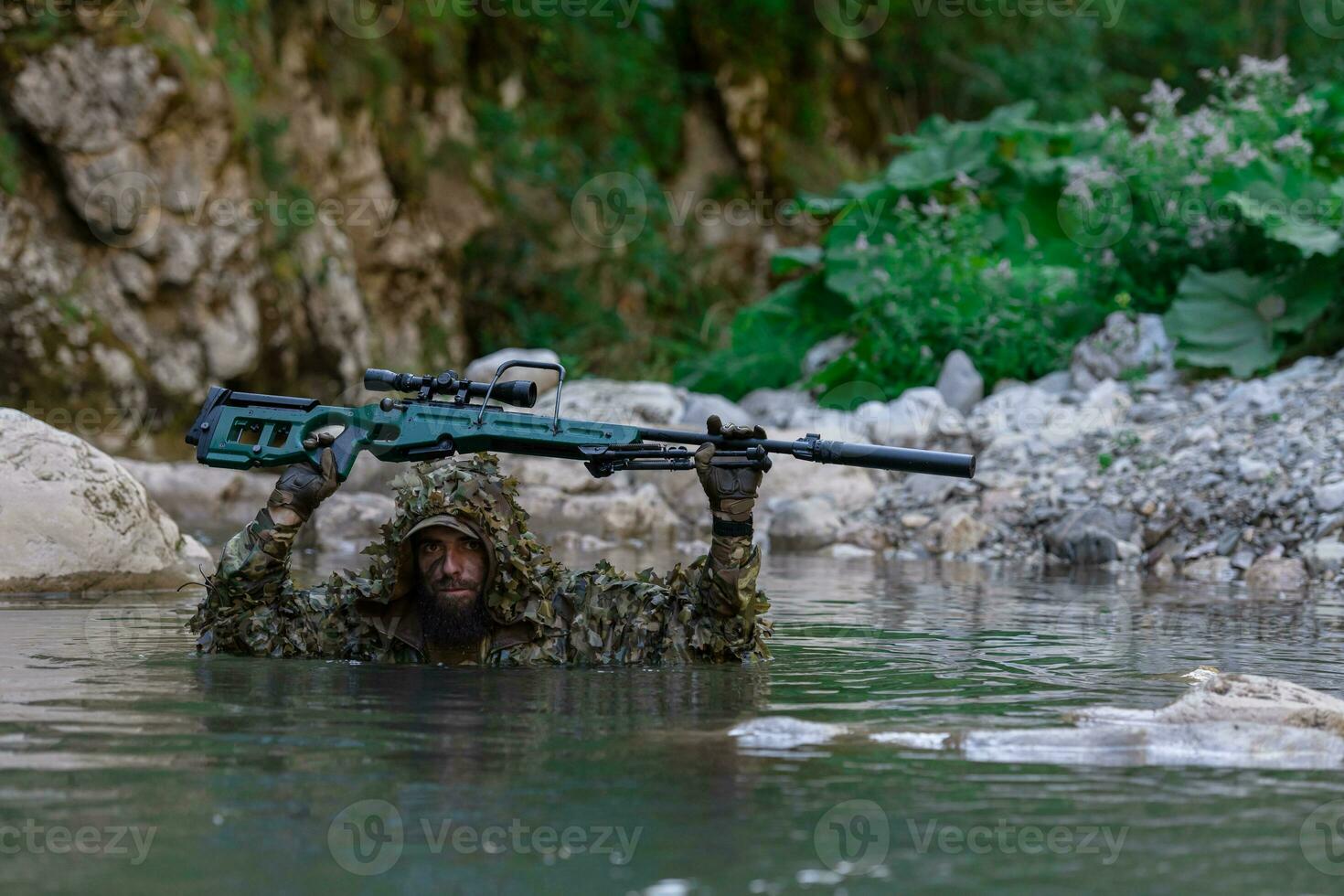 A military man or airsoft player in a camouflage suit sneaking the river and aims from a sniper rifle to the side or to target. photo