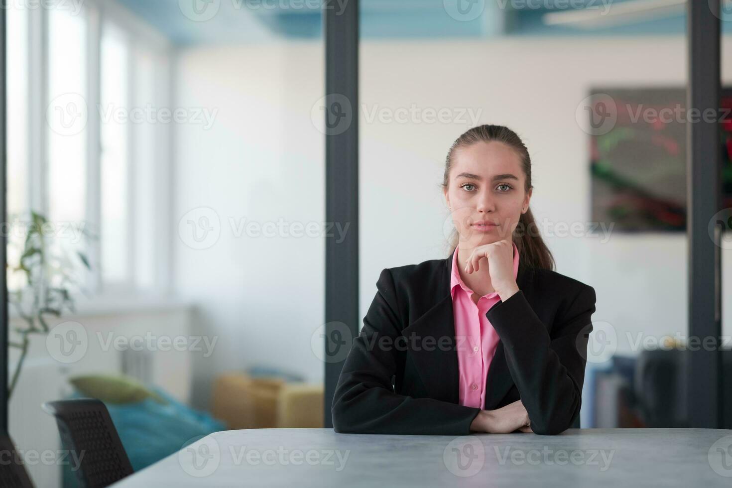 Successful young female leader in a suit with a pink shirt sitting in a modern glass office with a determined smile. photo