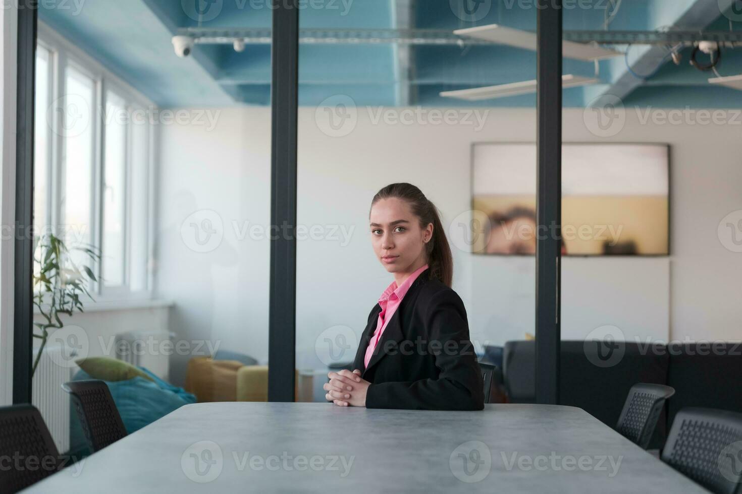 Successful young female leader in a suit with a pink shirt sitting in a modern glass office with a determined smile. photo