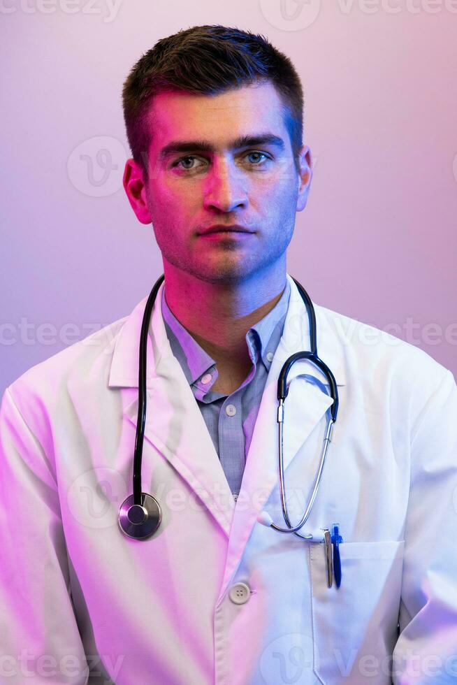 Coronavirus covid-19 danger alert Portrait of hero in white coat. Cheerful smiling young doctor with stethoscope in medical hospital standing against blue and pink background. photo