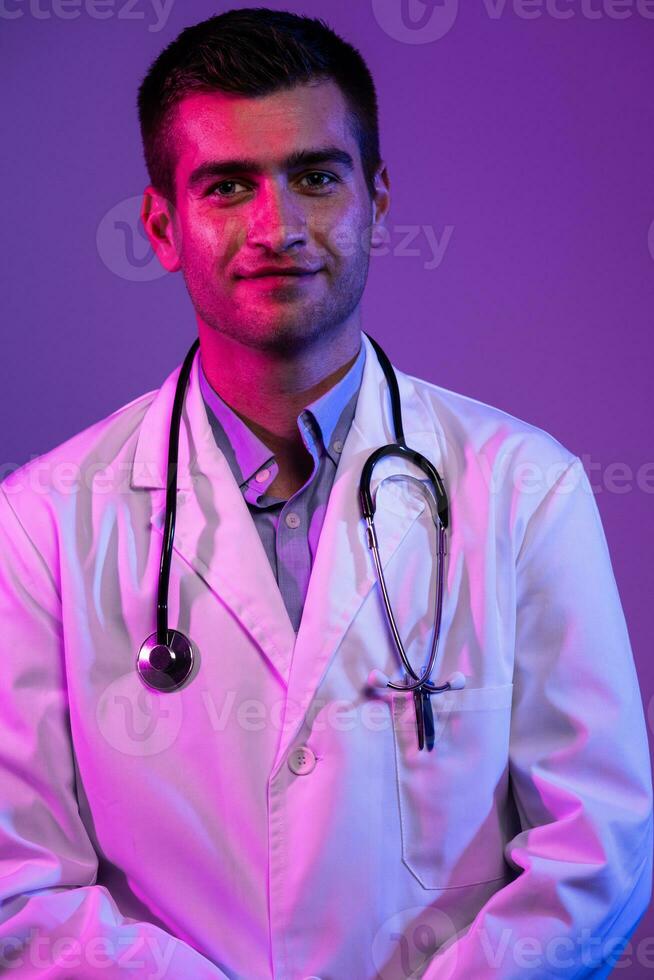 Coronavirus covid-19 danger alert Portrait of hero in white coat. Cheerful smiling young doctor with stethoscope in medical hospital standing against blue and pink background. photo