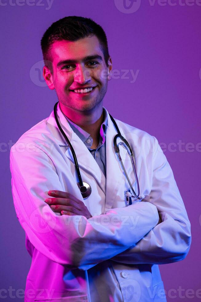 Portrait of hero in white coat. Cheerful smiling young doctor with stethoscope in medical hospital standing against blue and pink background. Coronavirus covid-19 danger alert photo