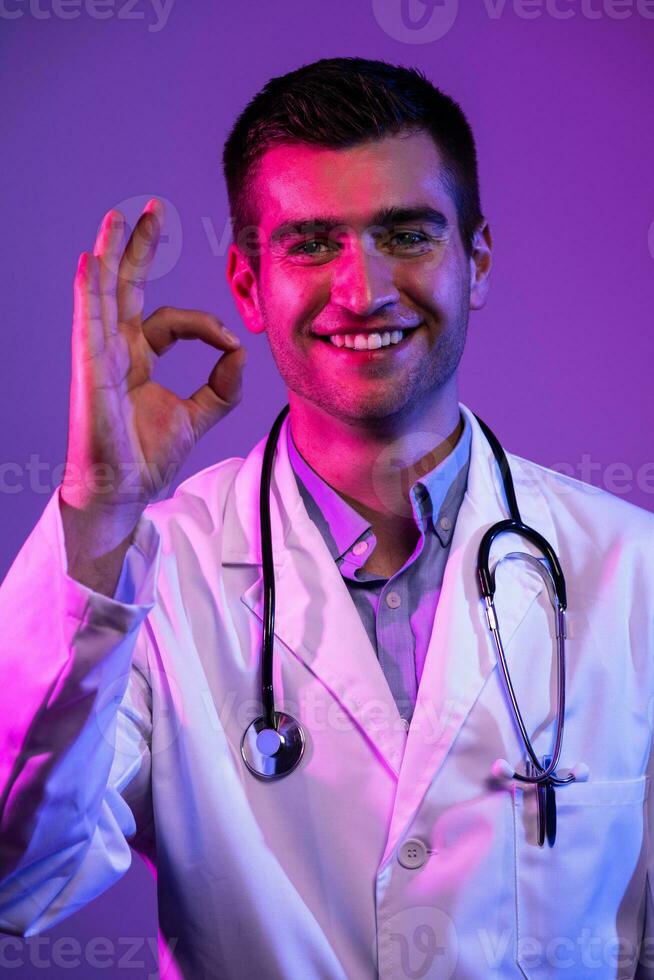 Doctor doing ok sign with fingers, excellent symbol neon lights blue and pink background. Coronavirus pandemic photo