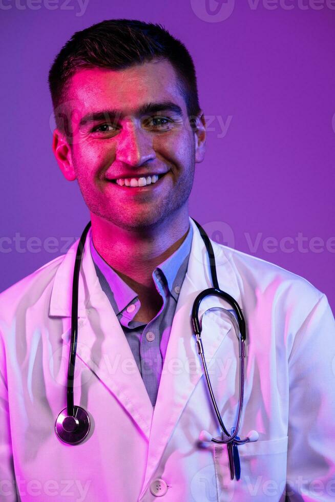 Coronavirus covid-19 danger alert Portrait of hero in white coat. Cheerful smiling young doctor with stethoscope in medical hospital standing against blue and pink background. photo