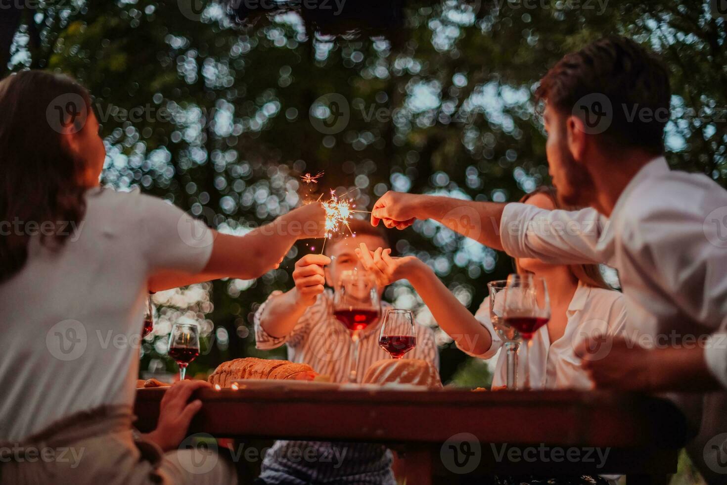 Group of happy friends celebrating holiday vacation using sprinklers and drinking red wine while having picnic french dinner party outdoor near the river on beautiful summer evening in nature photo