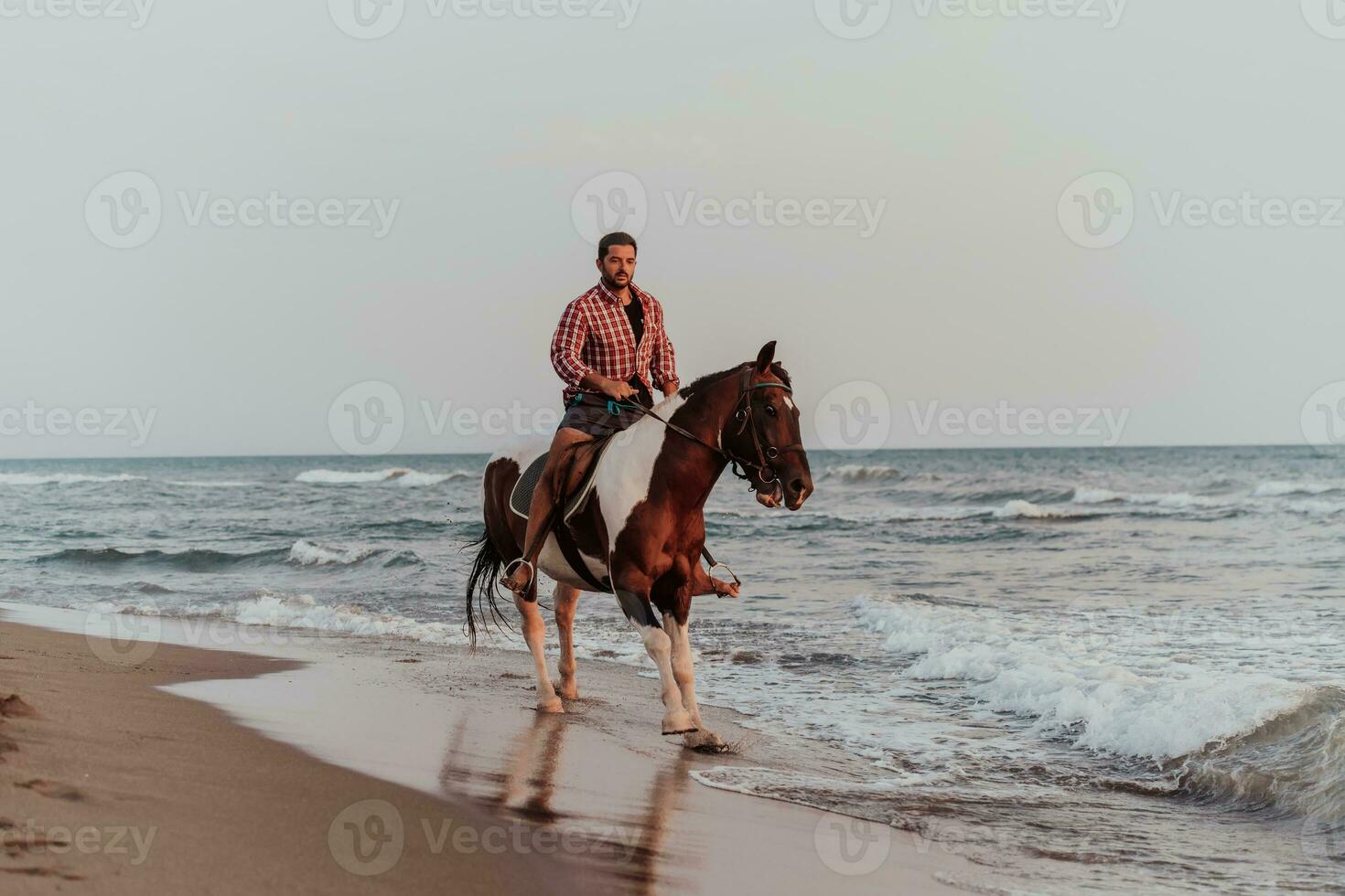 A modern man in summer clothes enjoys riding a horse on a beautiful sandy beach at sunset. Selective focus photo