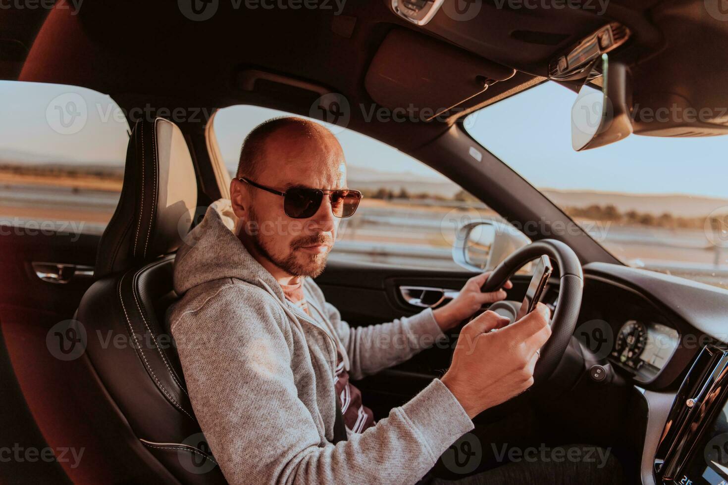 A man with a sunglasses driving a car and type a message on smartphone at sunset. The concept of car travel photo