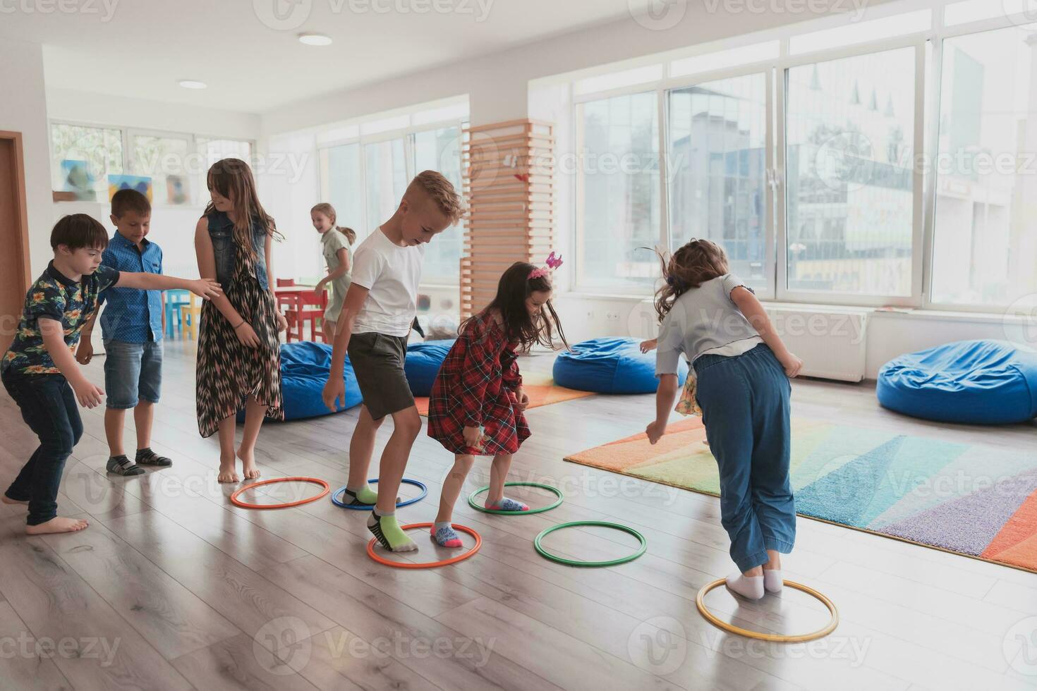 pequeño guardería colegio niños con hembra profesor en piso adentro en aula, haciendo ejercicio. saltando terminado hula aro círculos pista en el piso. foto