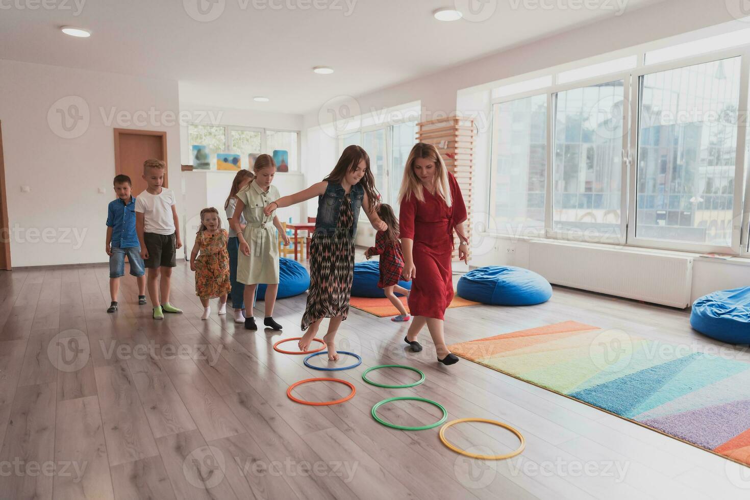 Small nursery school children with female teacher on floor indoors in classroom, doing exercise. Jumping over hula hoop circles track on the floor. photo