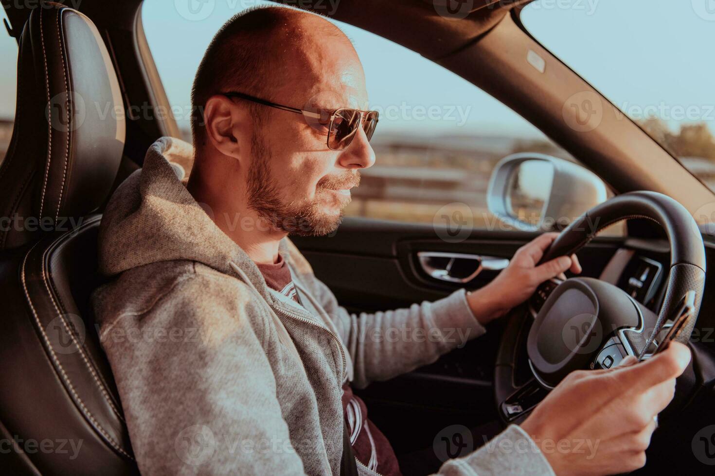 A man with a sunglasses driving a car and type a message on smartphone at sunset. The concept of car travel photo