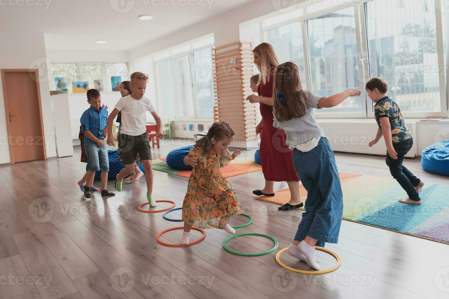 pequeño guardería colegio niños con hembra profesor en piso adentro en aula, haciendo ejercicio. saltando terminado hula aro círculos pista en el piso. foto