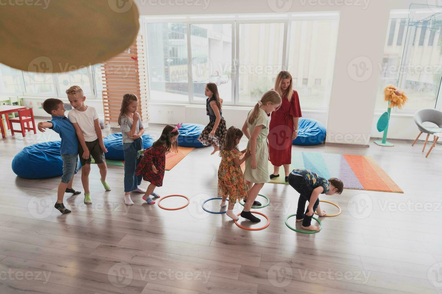 pequeño guardería colegio niños con hembra profesor en piso adentro en aula, haciendo ejercicio. saltando terminado hula aro círculos pista en el piso. foto