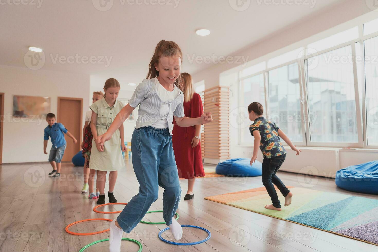 pequeño guardería colegio niños con hembra profesor en piso adentro en aula, haciendo ejercicio. saltando terminado hula aro círculos pista en el piso. foto