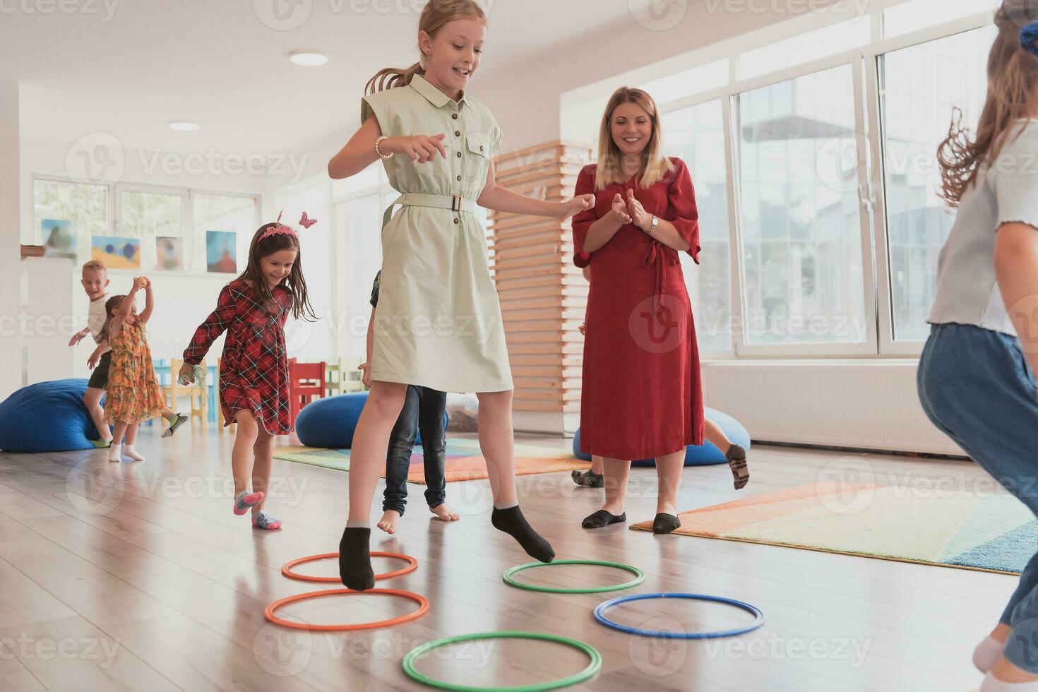 Small nursery school children with female teacher on floor indoors in classroom, doing exercise. Jumping over hula hoop circles track on the floor. photo