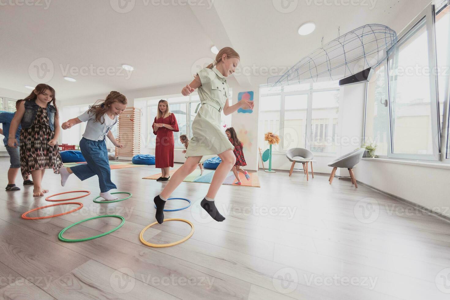 Small nursery school children with female teacher on floor indoors in classroom, doing exercise. Jumping over hula hoop circles track on the floor. photo