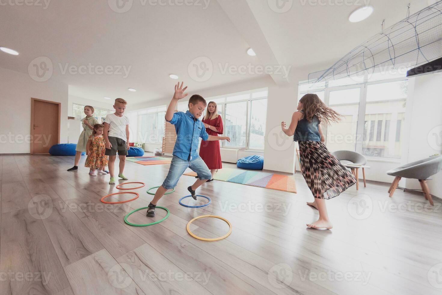 Small nursery school children with female teacher on floor indoors in classroom, doing exercise. Jumping over hula hoop circles track on the floor. photo