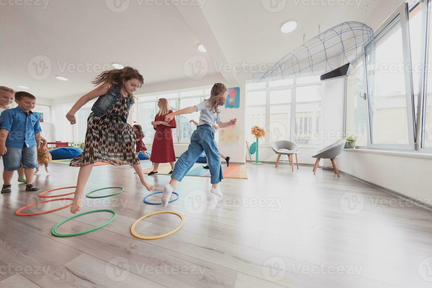 pequeño guardería colegio niños con hembra profesor en piso adentro en aula, haciendo ejercicio. saltando terminado hula aro círculos pista en el piso. foto