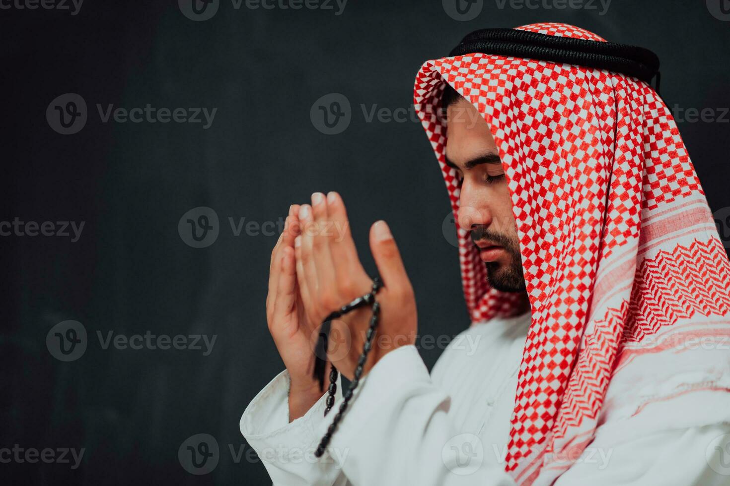 Arabian man in traditional clothes making traditional prayer to God, keeps hands in praying gesture in front of black chalkboard representing modern islam fashion and ramadan kareem concept photo