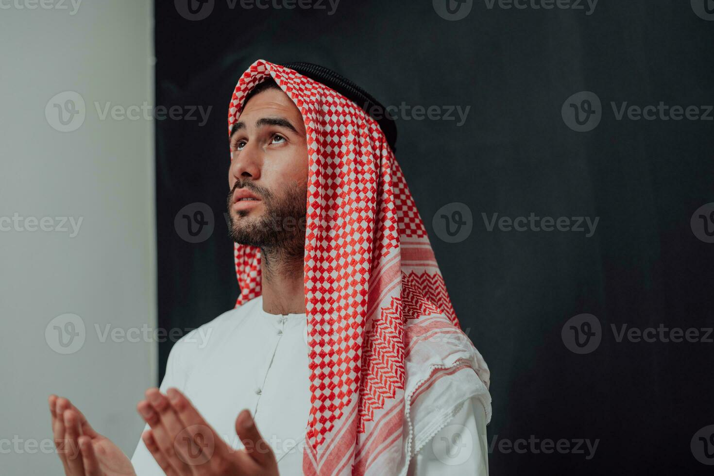 Arabian man in traditional clothes making traditional prayer to God, keeps hands in praying gesture in front of black chalkboard representing modern islam fashion and ramadan kareem concept photo