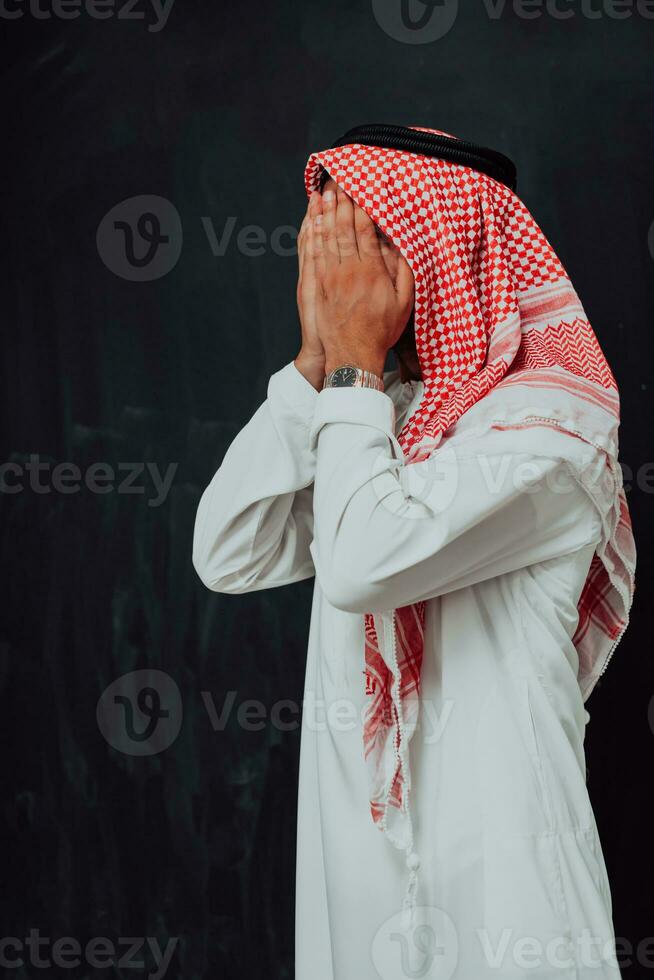 Arabian man in traditional clothes making traditional prayer to God, keeps hands in praying gesture in front of black chalkboard representing modern islam fashion and ramadan kareem concept photo