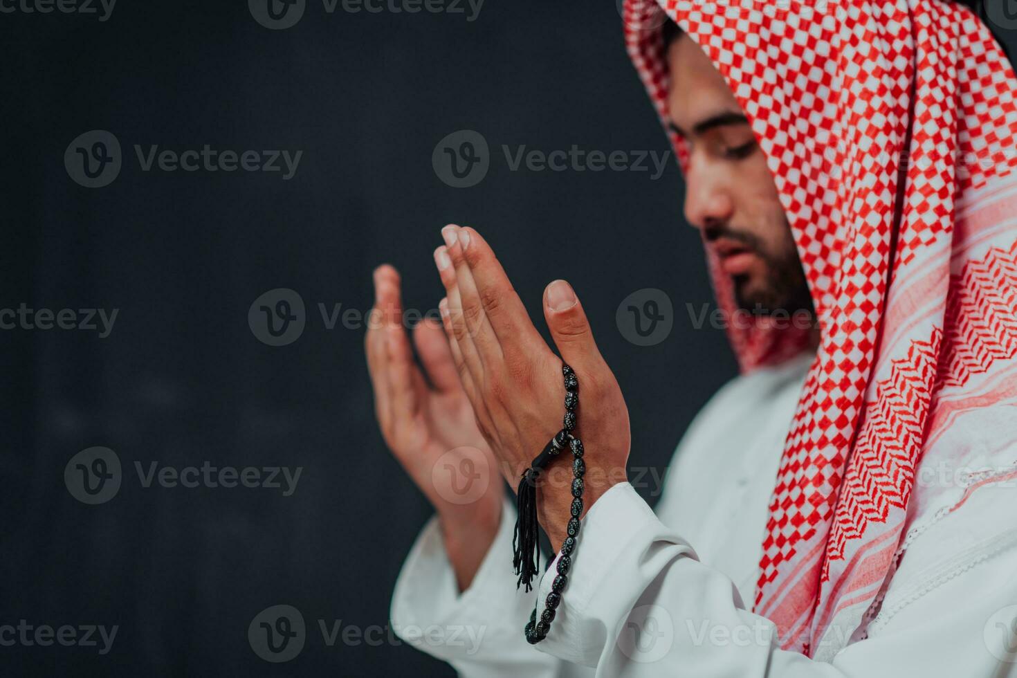 Arabian man in traditional clothes making traditional prayer to God, keeps hands in praying gesture in front of black chalkboard representing modern islam fashion and ramadan kareem concept photo