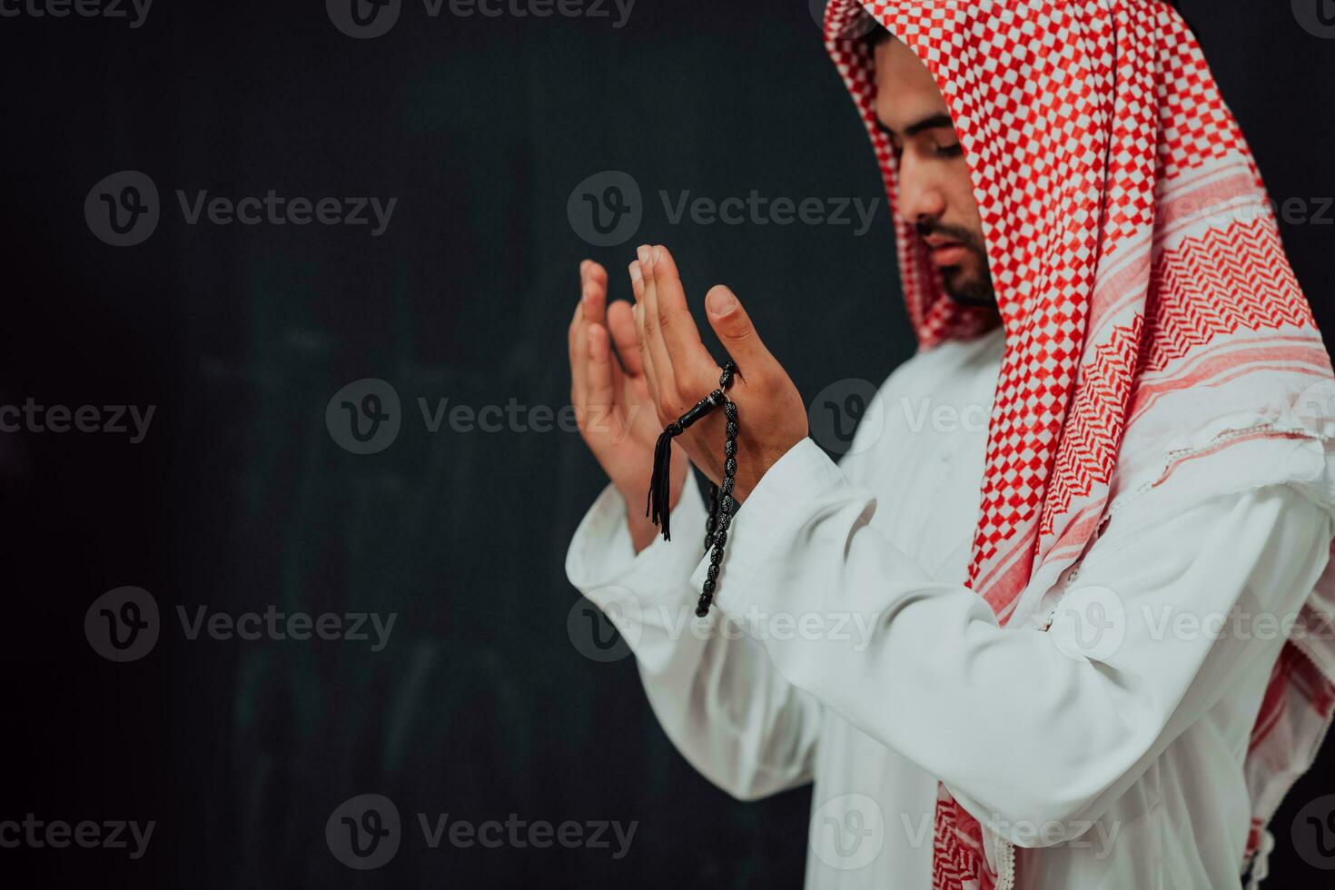 Arabian man in traditional clothes making traditional prayer to God, keeps hands in praying gesture in front of black chalkboard representing modern islam fashion and ramadan kareem concept photo
