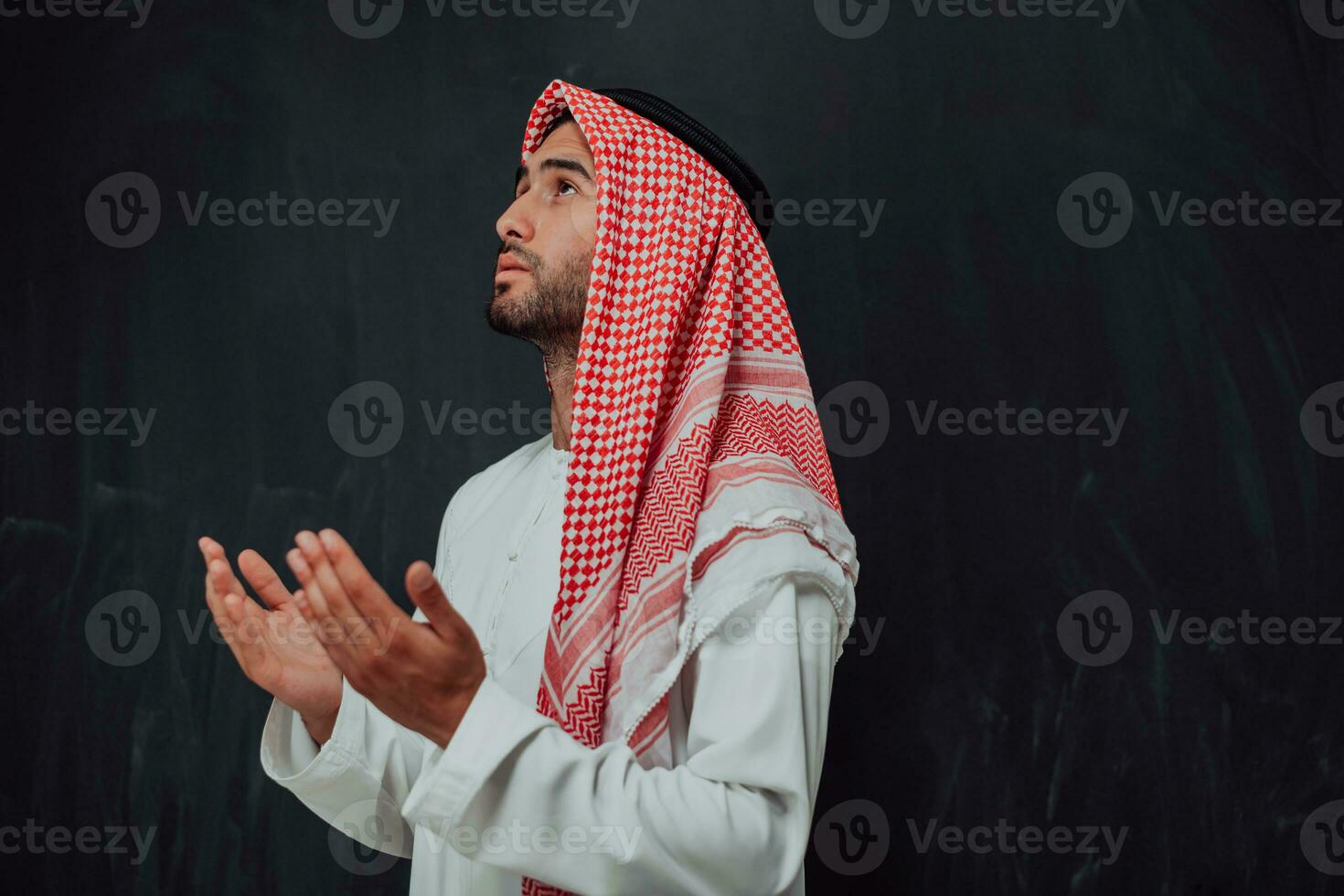 Arabian man in traditional clothes making traditional prayer to God, keeps hands in praying gesture in front of black chalkboard representing modern islam fashion and ramadan kareem concept photo