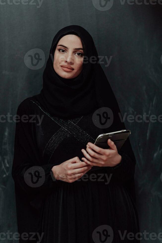 Young modern muslim business woman using tablet computer wearing glasses and hijab clothes in front of black chalkboard photo