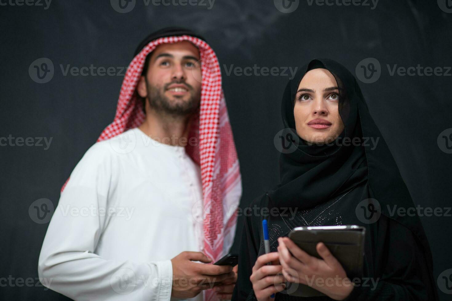 Young muslim business couple arabian man with woman in fashionable hijab dress using mobile phone and tablet computer in front of black chalkboard representing modern islam fashion technology photo