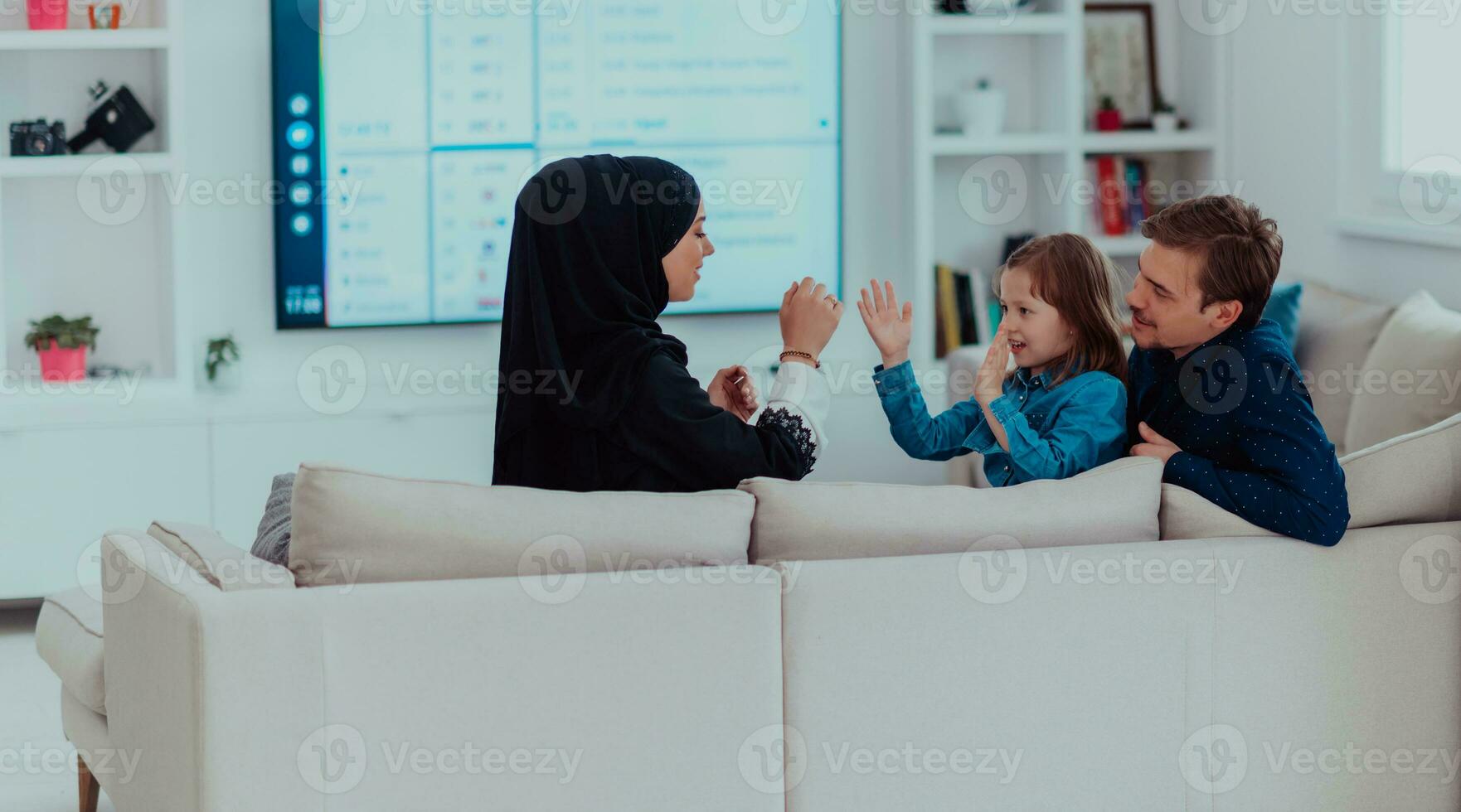 contento musulmán familia con hija mujer en tradicional de moda vestir teniendo divertido y bueno hora juntos mientras sentado en sofá foto