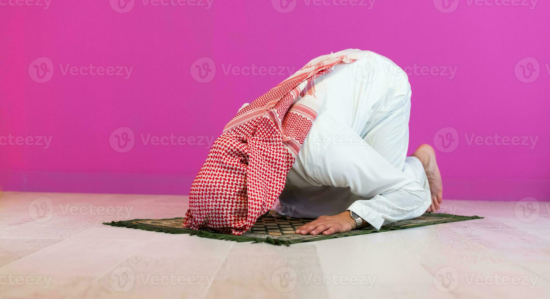Young arabian muslim man in traditional clothes praying on the carpet in front of pink wall before iftar dinner during a ramadan feast at home photo