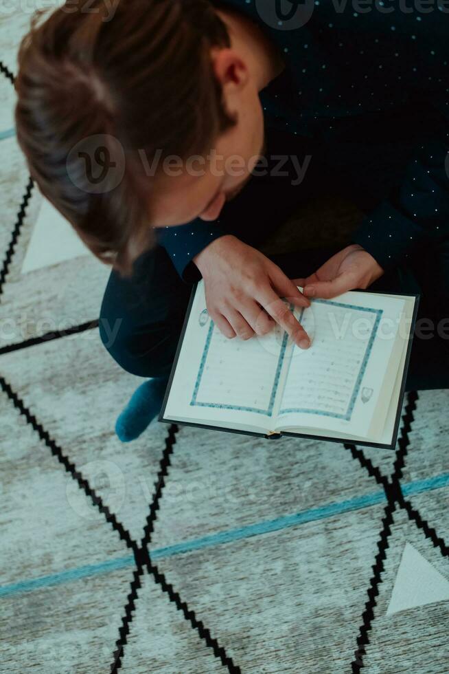 joven musulmán hombre sentado en el piso mientras leyendo santo libro Corán antes de iftar cena durante un Ramadán banquete a hogar foto