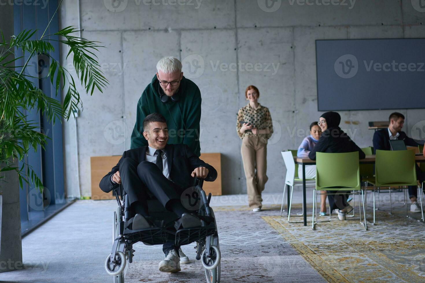 un joven empresario y su colega, quien es un persona con un discapacidad, divertido juntos mediante un grande, moderno oficina, demostrando el poder de trabajo en equipo, inclusión, y determinación como ellos conducir el silla de ruedas foto