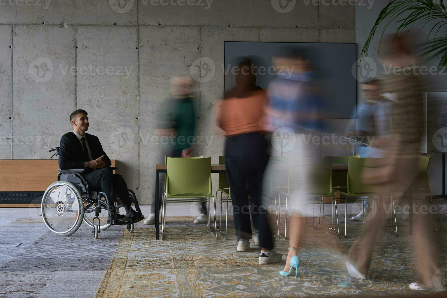 un empresario en un silla de ruedas en un moderno oficina, rodeado por su colegas quien son retratado con borroso movimientos, simbolizando su apoyo y solidaridad como ellos navegar el espacio de trabajo juntos. foto
