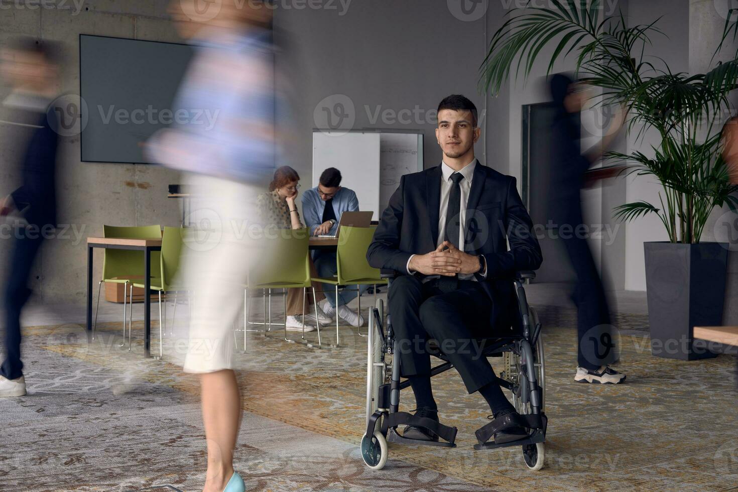 un empresario en un silla de ruedas en un moderno oficina, rodeado por su colegas quien son retratado con borroso movimientos, simbolizando su apoyo y solidaridad como ellos navegar el espacio de trabajo juntos. foto