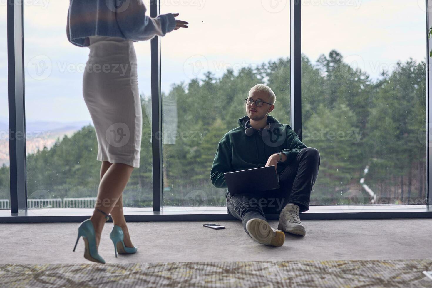 A young blond man in a modern office is sitting by the window, engrossed in his work on a laptop while talking to a female colleague photo