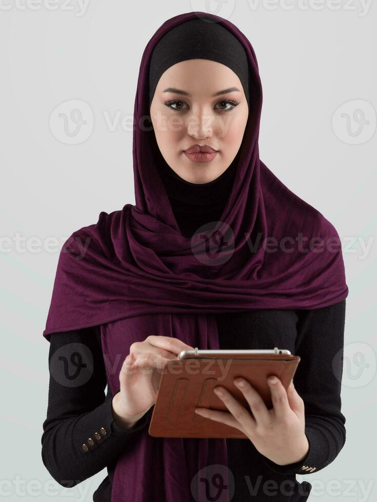 hermosa y moderno joven Arábica mujer de negocios en hijab utilizando un tableta computadora mientras posando en ligero antecedentes y sonriente a el cámara. negocio diversidad concepto, musulmán dama. foto