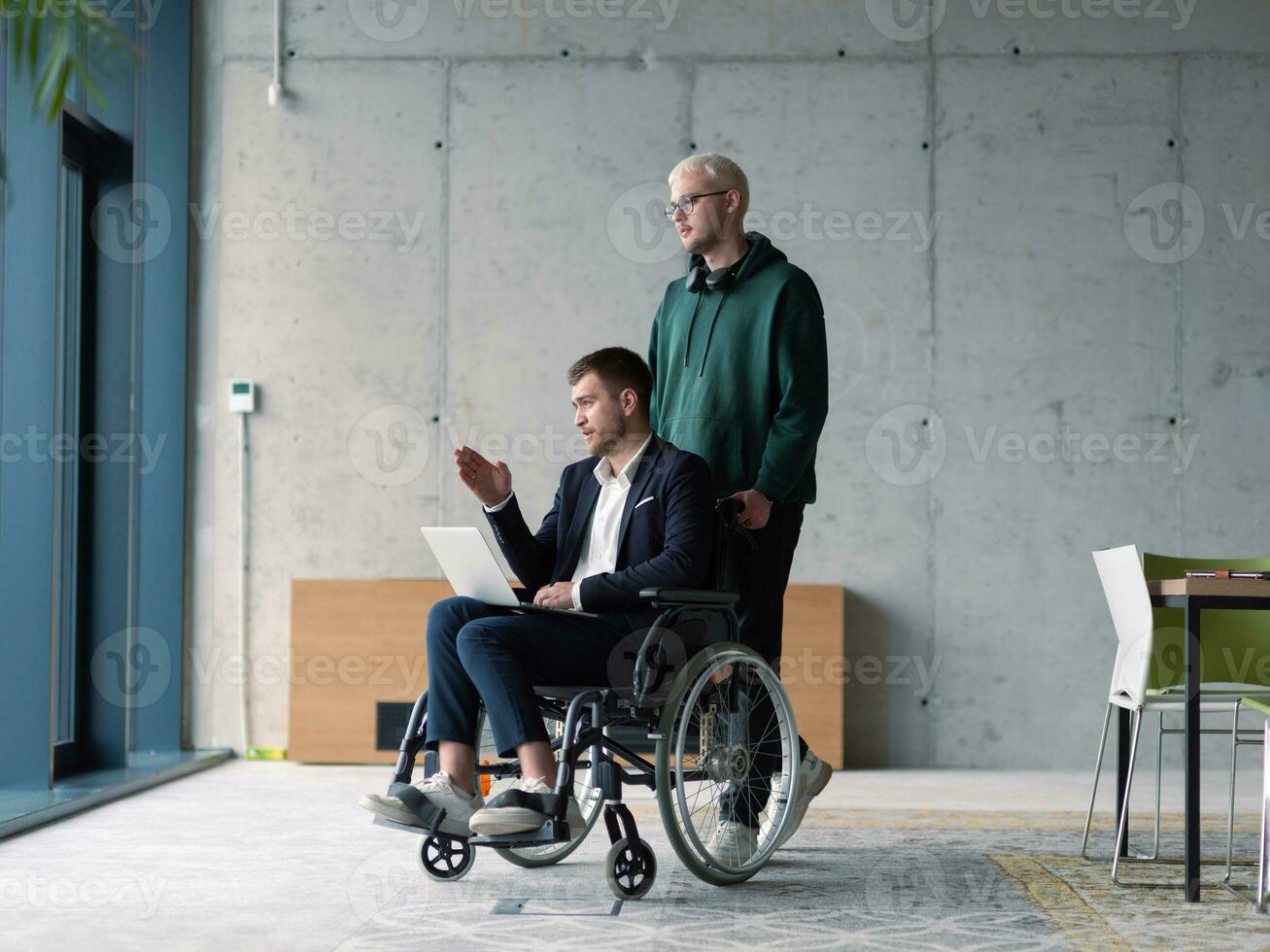A businessman in a wheelchair in a fashionable office using a laptop while behind him is his business colleague who gives him support photo