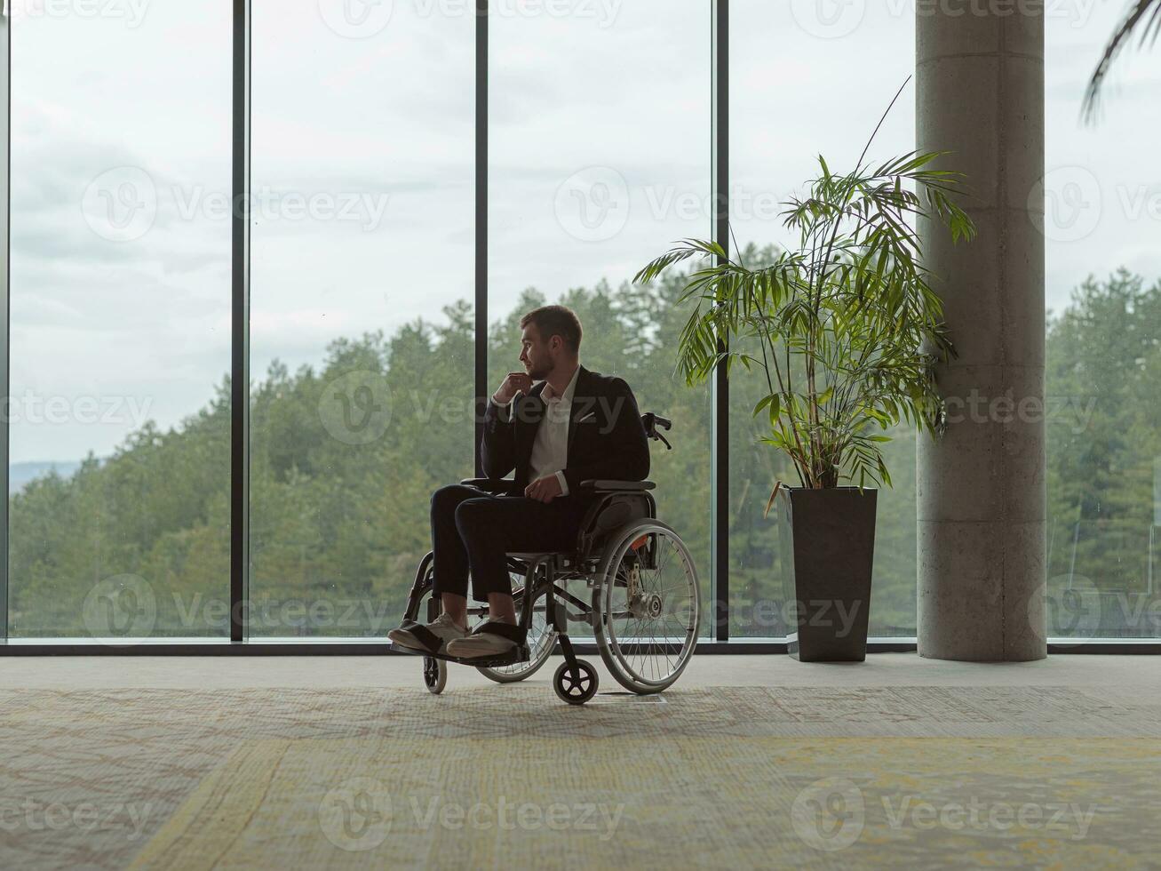 A melancholic businessman in a wheelchair sitting with a sad expression, gazing through the window of a modern office, conveying a sense of solitude photo